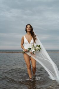 bride in bathing suit walking in water and holding lilys